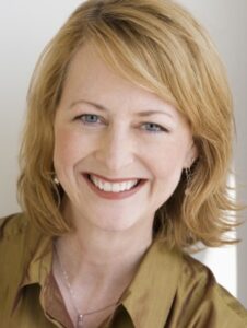 Smiling woman with shoulder-length blonde hair wearing a brown blouse, looking at the camera.