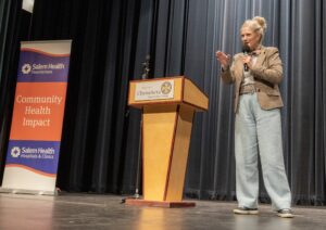 Speaker at a podium on stage, gesturing with one hand while holding a microphone; banners for Salem Health visible beside.