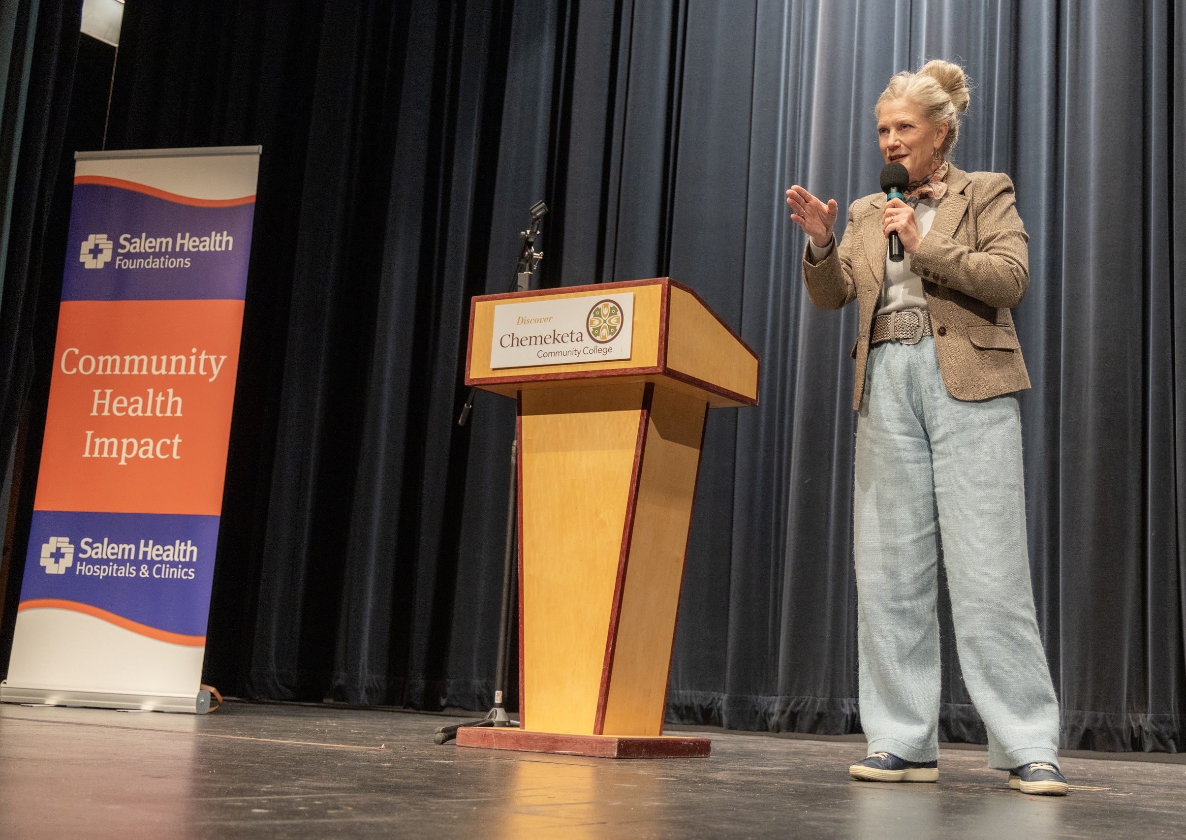 Speaker at a podium on stage, gesturing with one hand while holding a microphone; banners for Salem Health visible beside.